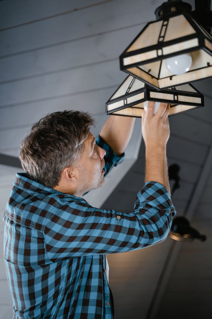 Home Adult male fixing pendant light indoors, wearing plaid long sleeves. Home maintenance scene.